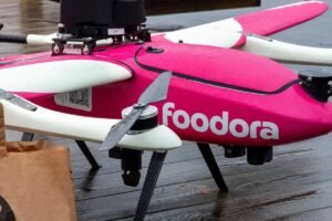 A imagem mostra um drone de entrega da Foodora, com um corpo predominantemente rosa. O drone possui quatro hélices e está posicionado sobre uma superfície de madeira. Ao lado do drone, há uma sacola de papel marrom, possivelmente contendo alimentos. O ambiente parece estar molhado, sugerindo que pode ter chovido recentemente.