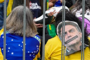 A imagem mostra duas pessoas de costas em um protesto. Uma delas está vestindo uma camiseta azul com a bandeira do Brasil, enquanto a outra usa uma camiseta amarela. A pessoa à direita tem um cartaz com a imagem de uma figura pública, com a palavra