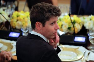 Um homem com cabelo castanho claro e liso, usando um terno escuro, está sentado em uma mesa de jantar. Ele tem uma expressão pensativa, com a mão no queixo. Ao fundo, há arranjos de flores amarelas e brancas, além de copos e pratos dispostos na mesa.