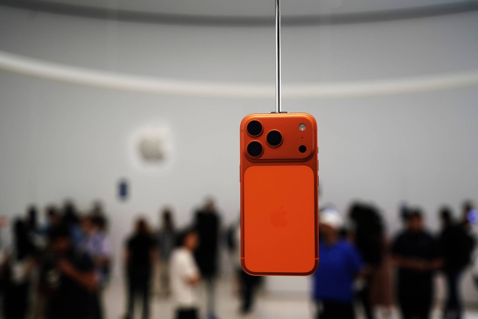 A imagem mostra um smartphone laranja pendurado em um suporte, com um fundo desfocado de pessoas em um evento. O smartphone possui três câmeras na parte traseira e o logotipo da Apple visível na parte inferior. O ambiente parece ser um espaço de apresentação, possivelmente durante um lançamento de produto.