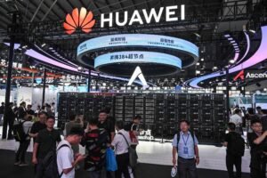 A imagem mostra um estande da Huawei em uma feira de tecnologia. No fundo, há um grande painel com o logotipo da Huawei e telas exibindo informações. Várias pessoas estão circulando pelo estande, observando os produtos expostos, que incluem servidores. O ambiente é bem iluminado e moderno, com uma grande quantidade de visitantes.