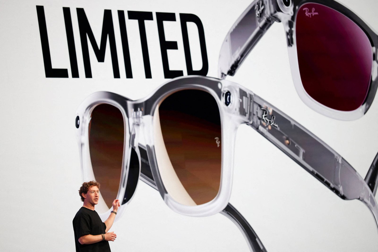 A imagem mostra um apresentador em pé à esquerda, gesticulando em direção a um grande painel de fundo. No painel, há uma imagem de óculos de sol Ray-Ban com armação transparente e lentes escuras. A palavra