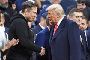 Elon Musk in a black hoodie shakes hands with Donald Trump wearing a navy coat and red striped tie. Several people stand closely behind them in a crowded indoor setting.