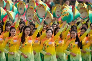 Várias mulheres vestindo áo dài amarelo com detalhes florais seguram chapéus cônicos decorados acima da cabeça em evento ao ar livre durante o dia.