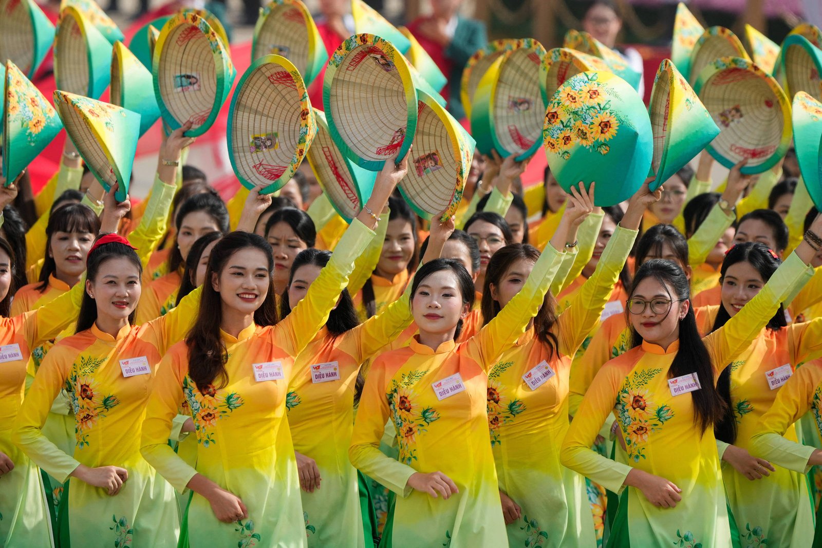 Várias mulheres vestindo áo dài amarelo com detalhes florais seguram chapéus cônicos decorados acima da cabeça em evento ao ar livre durante o dia.