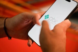 A imagem mostra uma pessoa segurando um smartphone com a tela exibindo o logotipo do aplicativo Pix. O fundo é desfocado, mas parece ser de uma superfície vermelha. A mão da pessoa está tocando a tela do celular.