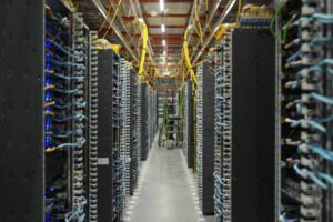 A imagem mostra uma sala de servidores com fileiras de equipamentos de computação. Os servidores estão organizados em racks altos, com cabos azuis e amarelos conectando-os. No fundo, há uma escada e uma pessoa em pé, possivelmente trabalhando nos equipamentos. A iluminação é artificial e o ambiente é bem organizado.