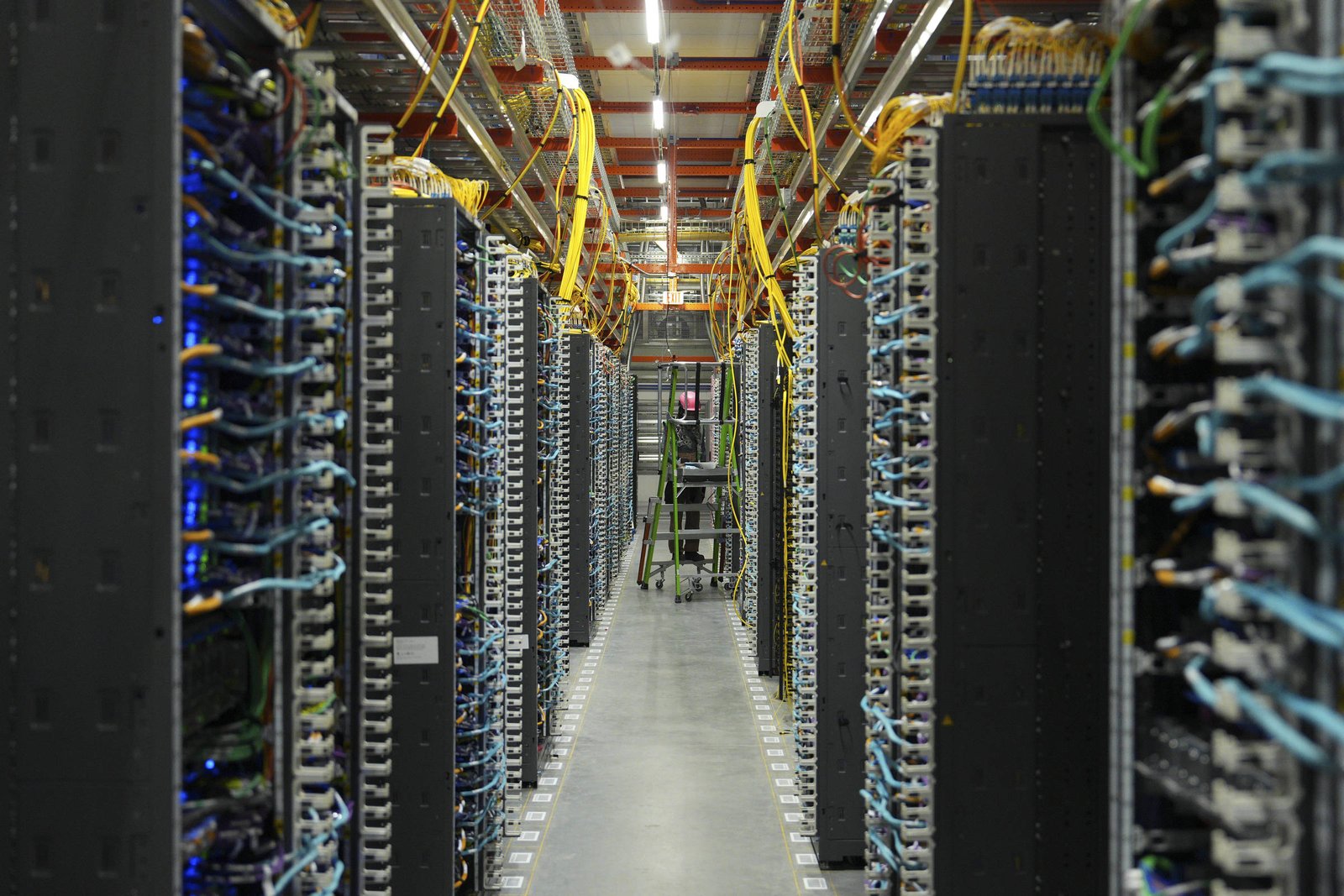 A imagem mostra uma sala de servidores com fileiras de equipamentos de computação. Os servidores estão organizados em racks altos, com cabos azuis e amarelos conectando-os. No fundo, há uma escada e uma pessoa em pé, possivelmente trabalhando nos equipamentos. A iluminação é artificial e o ambiente é bem organizado.