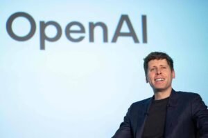 Um homem sorridente está sentado em uma cadeira, com um fundo que exibe o logotipo da OpenAI em letras grandes. Ele está vestindo uma jaqueta escura e parece estar em um evento ou apresentação.
