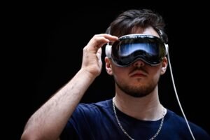 Homem jovem com barba curta e cabelo escuro usa óculos de realidade virtual pretos com detalhes brancos. Ele segura a lateral do óculos com a mão direita, veste camiseta azul escura e corrente prateada no pescoço. Fundo é totalmente preto.