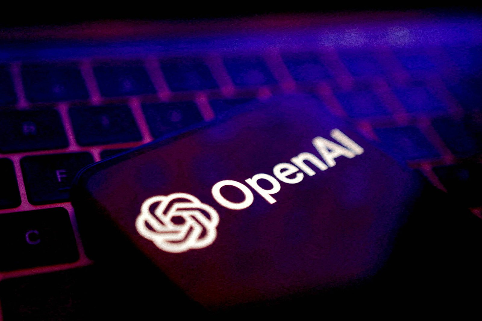 Logotipo branco da OpenAI exibido em tela escura de dispositivo eletrônico, posicionado sobre teclado iluminado por luz roxa.