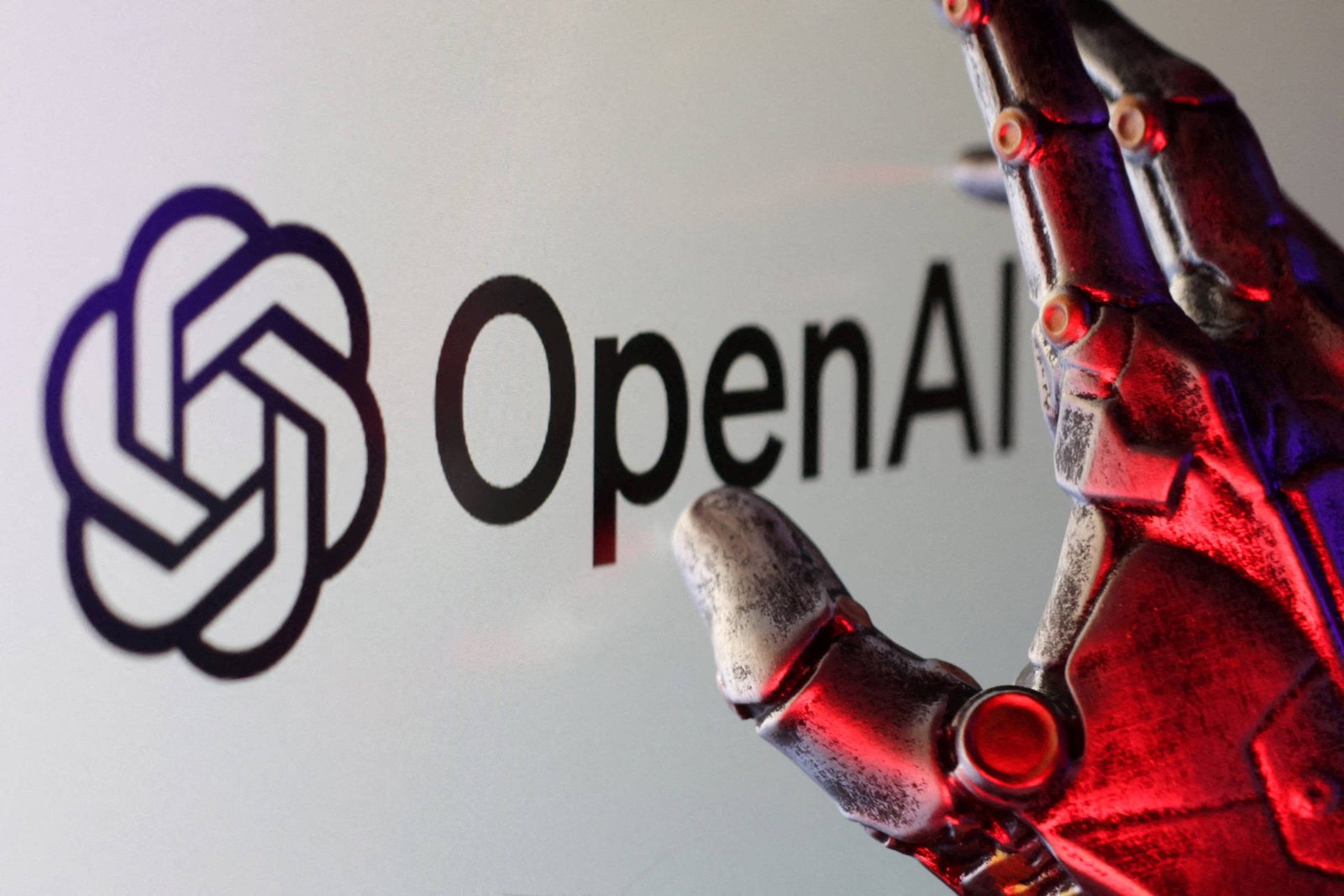 Mão metálica robótica iluminada por luz vermelha se aproxima do logotipo da OpenAI exibido em fundo claro.