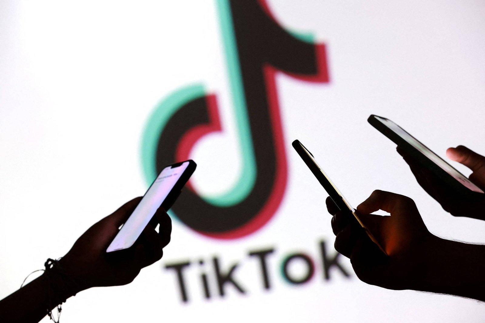 Três mãos seguram celulares com telas iluminadas em frente a uma tela branca exibindo o logo colorido do TikTok e o nome da plataforma em preto.
