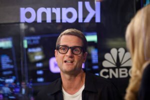 Homem de óculos e cabelo curto fala durante entrevista em estúdio com logotipos da Klarna e CNBC ao fundo.