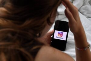 Pessoa de cabelos castanhos segura um celular com a tela exibindo o logo do Instagram. A mão direita está próxima à cabeça, e a pessoa está deitada sobre uma superfície branca.