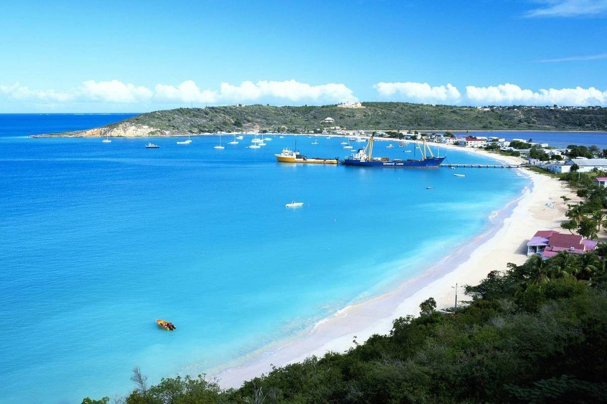 Vista panorâmica de praia com areia clara e mar azul turquesa. Ao fundo, porto com embarcações e cais, além de vegetação densa na margem direita. Céu azul com poucas nuvens.
