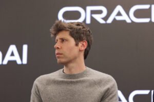 Homem jovem de cabelo castanho claro e curto, vestindo suéter cinza, olhando para o lado esquerdo. Fundo preto com logotipo branco da Oracle parcialmente visível.