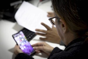 A imagem mostra uma pessoa de costas, usando óculos, focada em um smartphone que está em suas mãos. Ao fundo, há uma mão de outra pessoa apontando para um documento em uma mesa, que também possui um teclado visível. O ambiente parece ser um escritório.