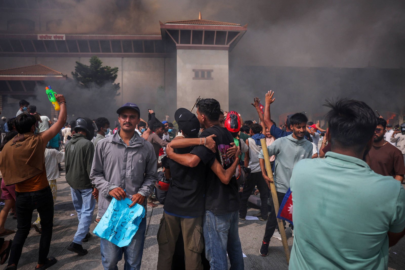 Multidão de manifestantes em praça com fumaça densa ao fundo. Pessoas erguem braços, algumas abraçadas, outras seguram cartazes e objetos. Edifício grande e cinza visível atrás da fumaça.