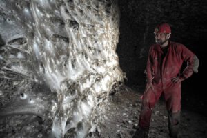Homem com macacão vermelho e capacete com lanterna observa formação mineral brilhante e texturizada em parede de caverna escura.