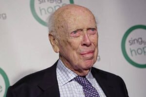 Homem idoso com terno preto, camisa xadrez branca e gravata roxa posando em frente a fundo branco com logotipo verde e texto