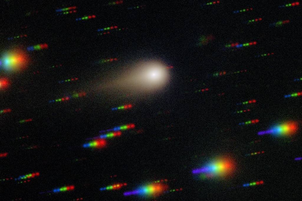 Cometa com núcleo brilhante e cauda difusa no centro da imagem, cercado por estrelas que aparecem como pontos coloridos em vermelho, verde e azul devido ao efeito de dispersão cromática. Fundo escuro do espaço.