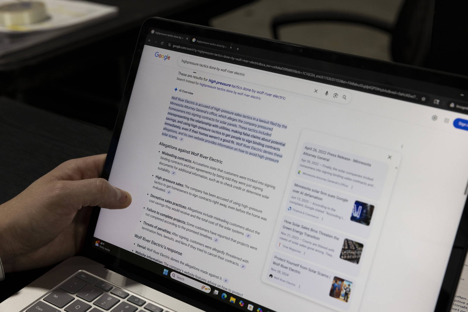 Mão esquerda segurando laptop aberto com tela exibindo resultados de pesquisa do Google sobre energia nuclear. Ambiente interno com mesa preta e cadeira ao fundo.