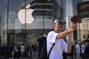 Jovem com mochila e camiseta branca usa óculos de realidade virtual e segura smartphone com estabilizador em frente à fachada de vidro da loja Apple, com o logo da maçã visível. Pessoas circulam ao fundo na calçada.