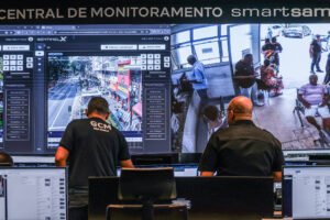 Dois operadores da Central de Monitoramento Smart Sampa observam telas grandes com imagens de câmeras de segurança mostrando uma rua movimentada e uma sala com várias pessoas sentadas e em pé. Eles estão sentados em frente a vários monitores menores com dados e imagens.