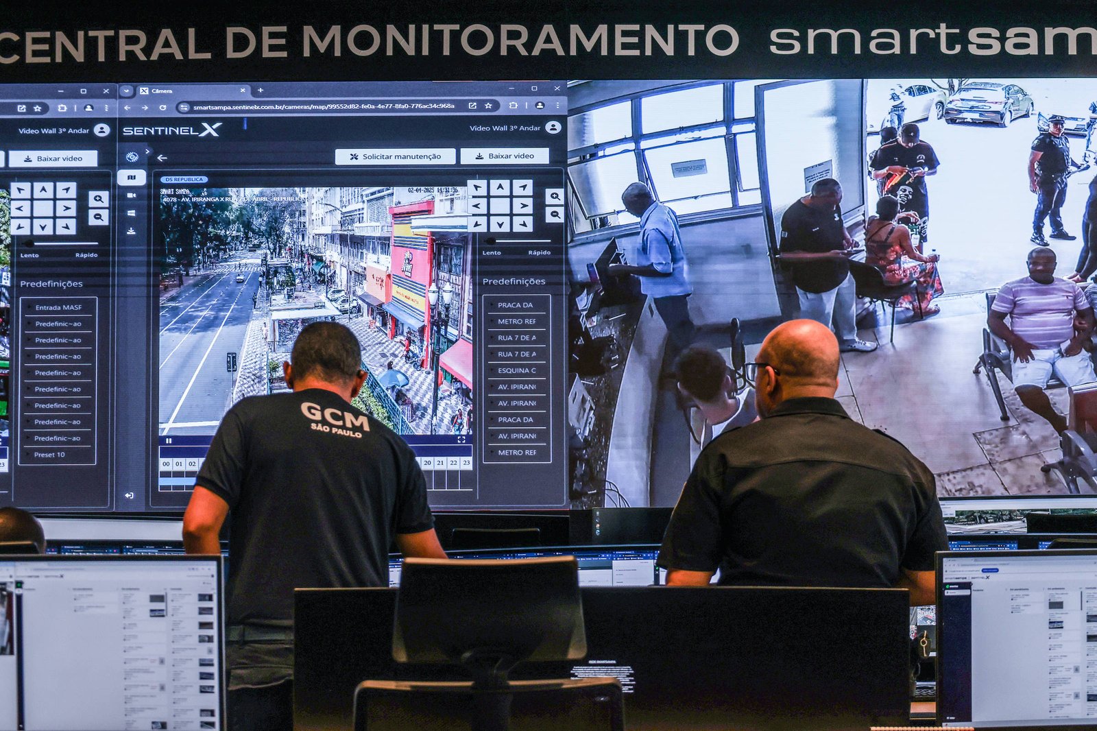 Dois operadores da Central de Monitoramento Smart Sampa observam telas grandes com imagens de câmeras de segurança mostrando uma rua movimentada e uma sala com várias pessoas sentadas e em pé. Eles estão sentados em frente a vários monitores menores com dados e imagens.