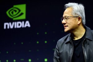 Homem de cabelos grisalhos e óculos, vestindo jaqueta preta, olha para a direita em frente a tela com o logotipo verde e branco da Nvidia.