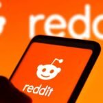 Pessoa segurando celular com logomarca do Reddit na tela e, ao fundo, também aparece a logomarca do Reddit