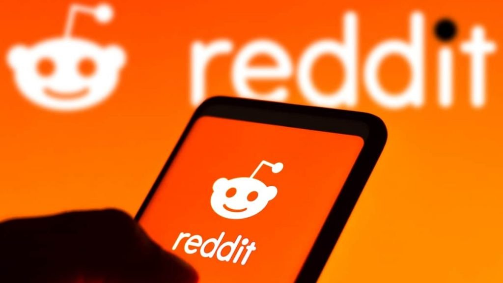 Pessoa segurando celular com logomarca do Reddit na tela e, ao fundo, também aparece a logomarca do Reddit