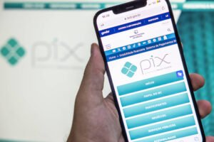 A imagem mostra uma mão segurando um smartphone que exibe a interface do aplicativo Pix. Na tela do celular, é possível ver o logotipo do Pix e opções como