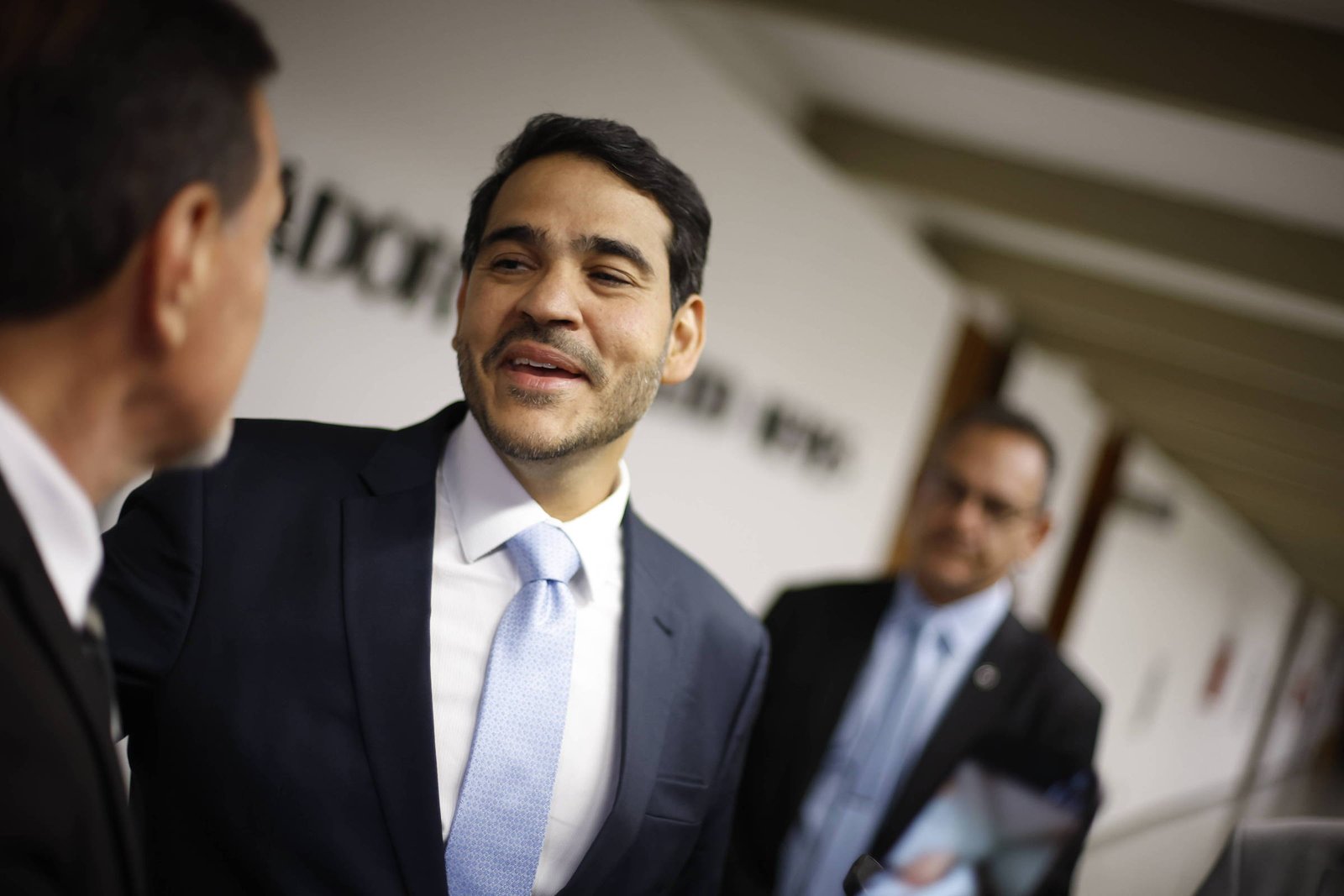 Três homens vestidos com ternos escuros e gravatas estão em um corredor com paredes brancas e teto com vigas expostas. Dois homens estão em primeiro plano conversando, enquanto o terceiro está ao fundo encostado na parede, observando. O ambiente sugere um local formal, possivelmente governamental ou corporativo.