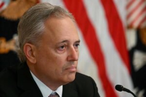 Homem de meia-idade com cabelo grisalho veste terno preto e gravata, sentado diante de microfone. Ao fundo, bandeiras dos Estados Unidos desfocadas.