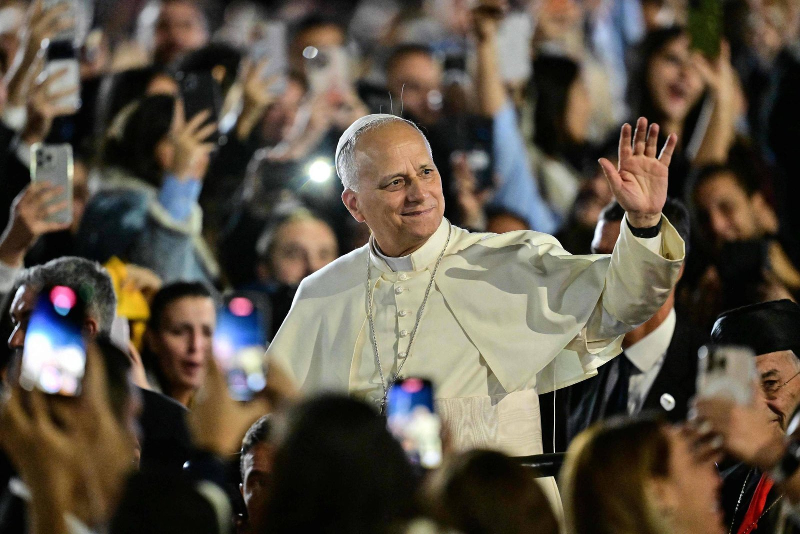 Líder religioso vestido com vestes brancas acena com a mão direita para uma multidão ao redor. Pessoas ao fundo seguram celulares para registrar o momento, em ambiente externo e iluminado.