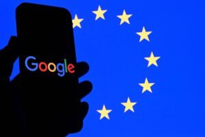 Mão segurando celular com tela preta exibindo o logotipo colorido do Google. Ao fundo, bandeira azul da União Europeia com doze estrelas amarelas dispostas em círculo.
