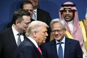 Grupo de homens em trajes formais, incluindo ternos ocidentais e vestimentas tradicionais árabes, posam juntos em ambiente interno com fundo azul. Entre eles, um homem de cabelo branco e terno escuro conversa com outro de terno e gravata, enquanto outros observam ao redor.