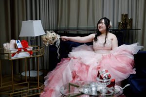 Jovem mulher sentada em sofá azul escuro, vestindo vestido volumoso rosa com camadas de tule. Ela sorri e estende o braço segurando um buquê de flores brancas. Ao lado, mesa de vidro com objetos decorativos e carrinho dourado com presentes e arranjo floral. Cortinas bege ao fundo e luminárias branca e de chão completam o ambiente interno.