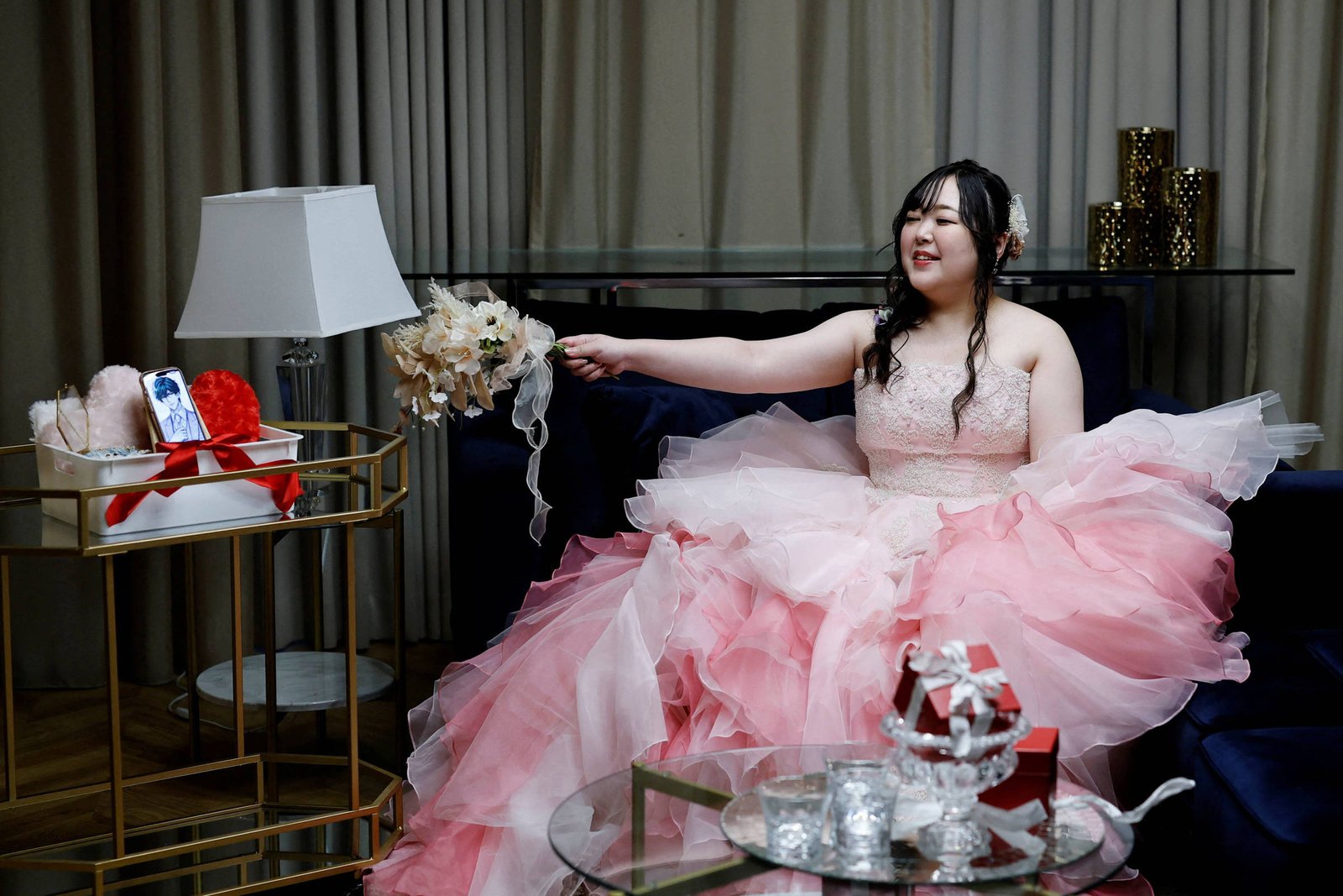 Jovem mulher sentada em sofá azul escuro, vestindo vestido volumoso rosa com camadas de tule. Ela sorri e estende o braço segurando um buquê de flores brancas. Ao lado, mesa de vidro com objetos decorativos e carrinho dourado com presentes e arranjo floral. Cortinas bege ao fundo e luminárias branca e de chão completam o ambiente interno.