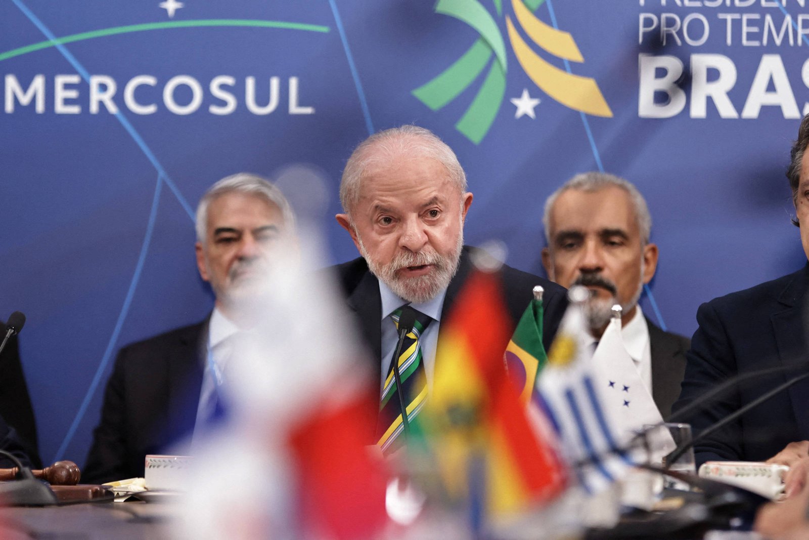 Homem de cabelos grisalhos e barba branca fala ao microfone em mesa com bandeiras de países do Mercosul. Ao fundo, painel azul com logotipo do Mercosul e texto indicando presidência pro tempore do Brasil. Outras três pessoas estão sentadas atrás dele.
