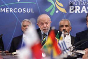 Homem de cabelos grisalhos e barba branca fala ao microfone em mesa com bandeiras de países do Mercosul. Ao fundo, painel azul com logotipo do Mercosul e texto indicando presidência pro tempore do Brasil. Outras três pessoas estão sentadas atrás dele