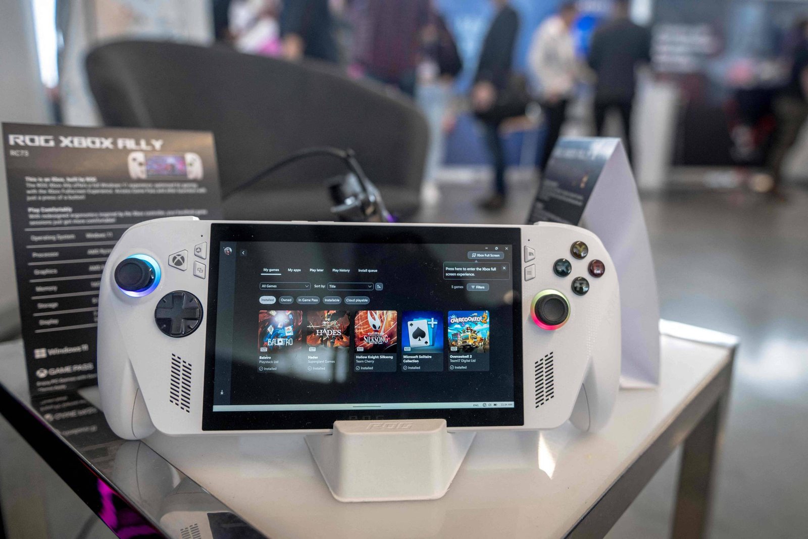 Console portátil branco ROG Xbox Ally exibido em suporte sobre mesa. Tela mostra interface de jogos disponíveis. Fundo desfocado com pessoas em ambiente de evento.
