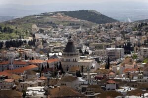 Vista panorâmica da cidade de Nazaré, com a Basílica da Anunciação em destaque no centro, cercada por edifícios residenciais e comerciais de telhados vermelhos. Ao fundo, colinas verdes e áreas urbanas se estendem sob céu claro.