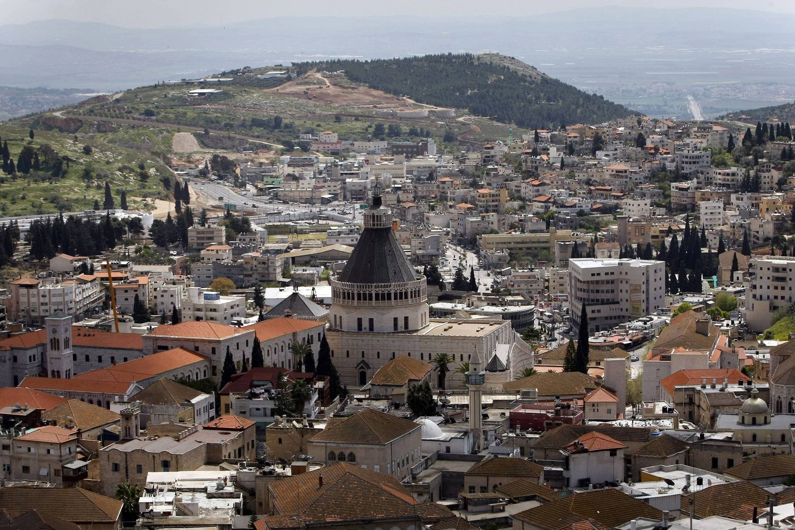Vista panorâmica da cidade de Nazaré, com a Basílica da Anunciação em destaque no centro, cercada por edifícios residenciais e comerciais de telhados vermelhos. Ao fundo, colinas verdes e áreas urbanas se estendem sob céu claro.