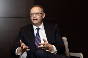 Homem de meia-idade veste terno escuro, camisa branca e gravata listrada, sentado em poltrona clara contra fundo escuro, gesticulando com as mãos enquanto fala.