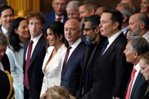 Grupo de executivos e empresários alinhados em ambiente interno, todos vestidos com trajes formais. Entre eles, destacam-se Jeff Bezos, Sundar Pichai e Elon Musk, posicionados no centro da imagem. O cenário sugere uma cerimônia ou reunião oficial, com pessoas ao fundo e iluminação quente.