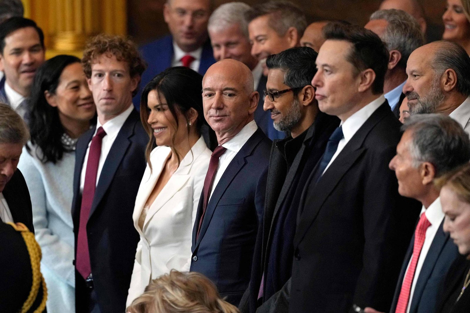 Grupo de executivos e empresários alinhados em ambiente interno, todos vestidos com trajes formais. Entre eles, destacam-se Jeff Bezos, Sundar Pichai e Elon Musk, posicionados no centro da imagem. O cenário sugere uma cerimônia ou reunião oficial, com pessoas ao fundo e iluminação quente.