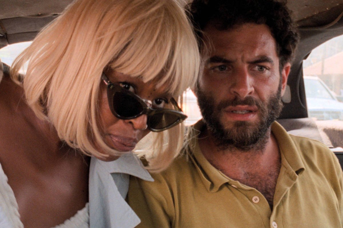 Uma mulher de cabelo loiro curto e óculos escuros está inclinada para frente ao lado de um homem de barba e cabelo escuro, que veste uma camisa amarela. Ambos estão no banco traseiro de um veículo, com sacolas visíveis ao lado direito.